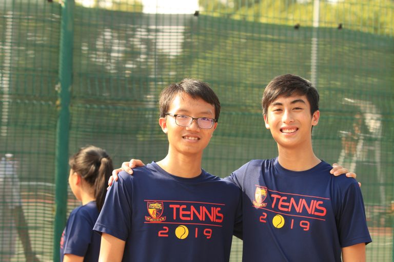 Two ACS(I) tennis team members in Tennis 2019 shirts smile together for a photo at a tennis event.